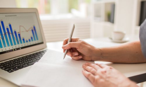 Close-up shot of busy manager taking necessary notes while analyzing statistics with help of modern laptop, blurred background